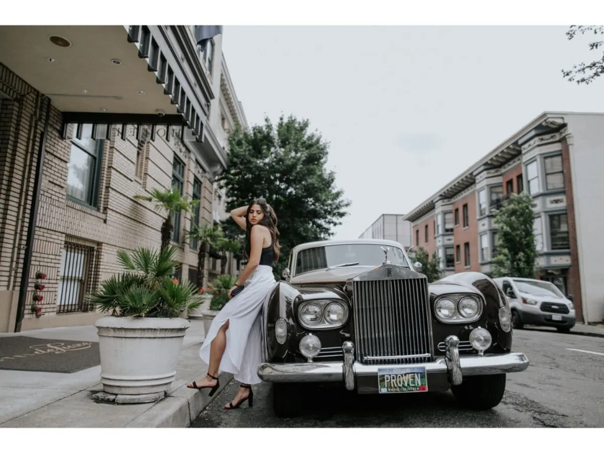 This stylish exterior shot features a classic car parked in front of the hotel's ornate urban architecture. A woman in a white dress stands nearby, surrounded by potted plants, evoking a sense of vintage glamour and inviting sophistication. The image captures the lively city atmosphere and the promise of memorable strolls just outside the hotel doors.