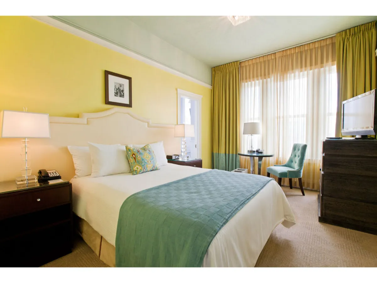 The image showcases a stylish Premium King room with a plush bed draped in a teal throw blanket, bathed in natural light from oversized windows adorned with sheer curtains. A small round table and chair sit by the window, inviting guests to relax, read, or enjoy a morning cup of tea while overlooking the cityscape. The space is warm, comfortable, and elegantly appointed, reflecting the boutique hotel's blend of classic and modern style.