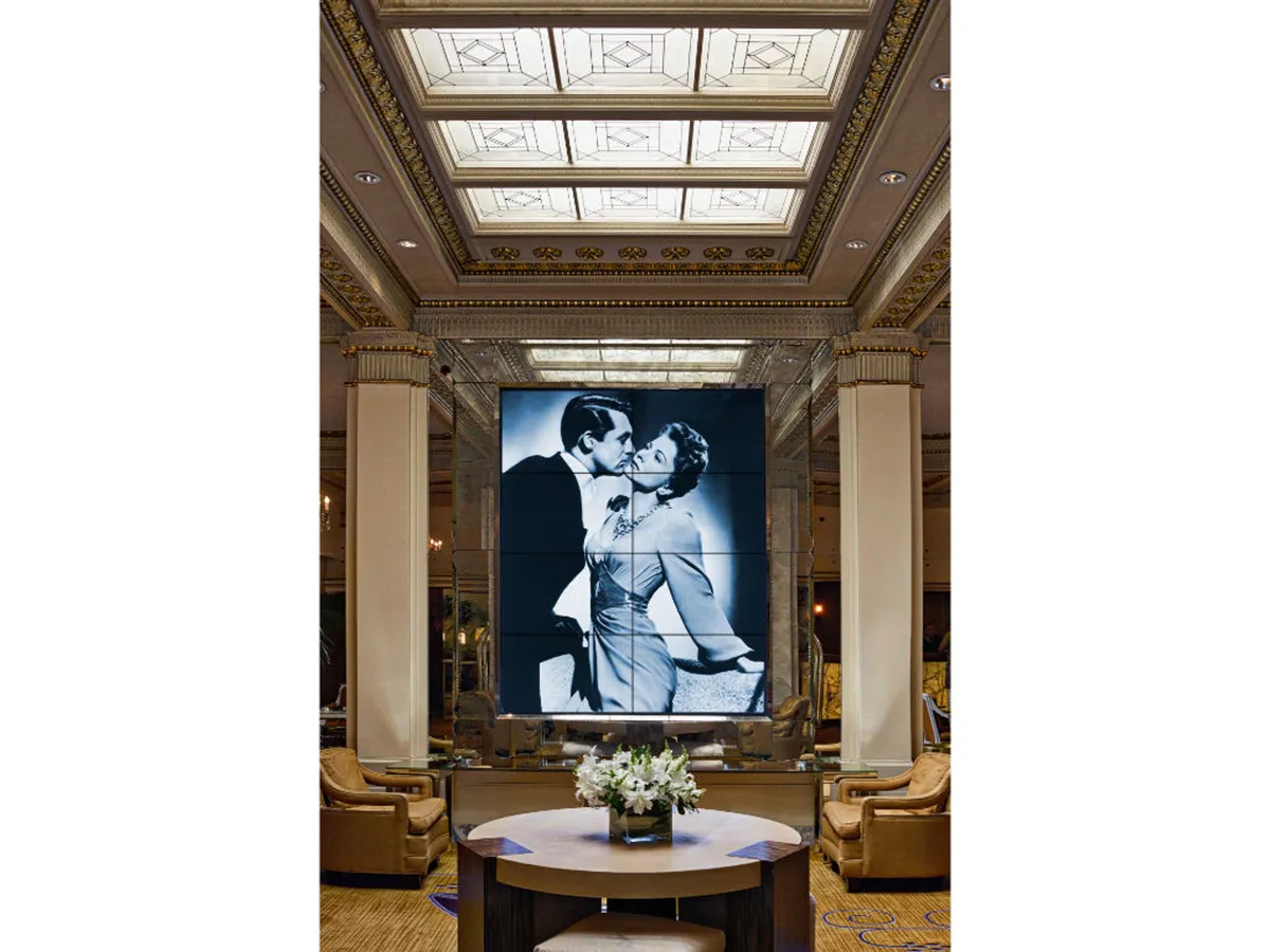 This image features a stylish interior space with a large screen prominently displaying a classic black and white romantic film image, surrounded by a central round table adorned with a floral arrangement and luxurious armchairs. The atmosphere is set for gatherings and film appreciation in a setting that evokes nostalgia and romance.