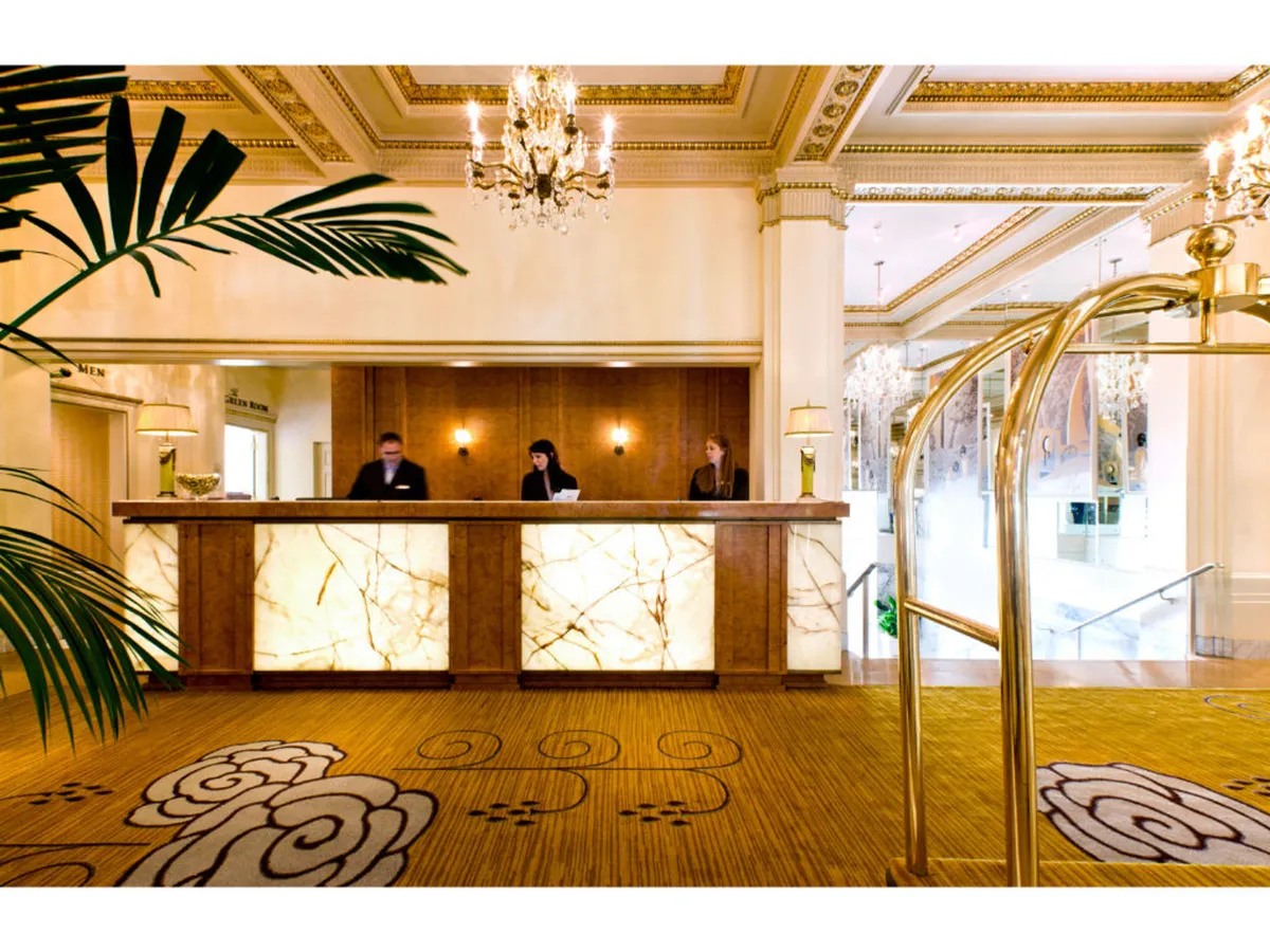 A luxurious hotel lobby welcomes guests, with a grand reception desk staffed by attentive employees. The space features a beautiful chandelier overhead and a plush floral carpet, setting an elegant and inviting tone for arrivals and check-ins.