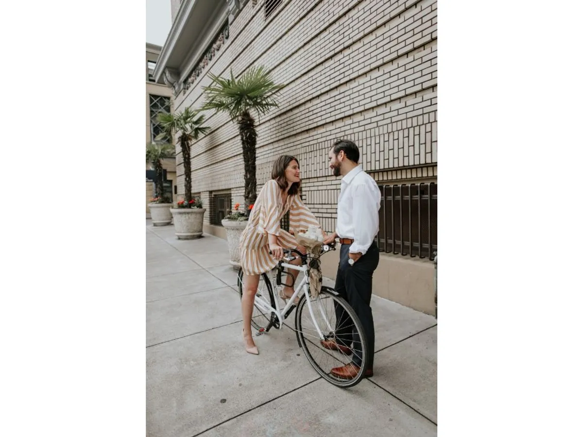 This exterior image captures a couple engaging in a lighthearted conversation beside a bicycle, set against a charming brick wall with potted plants. The bicycle subtly references the hotel's complimentary bike rentals, while the couple's relaxed demeanor evokes the playful and leisurely spirit of urban exploration.