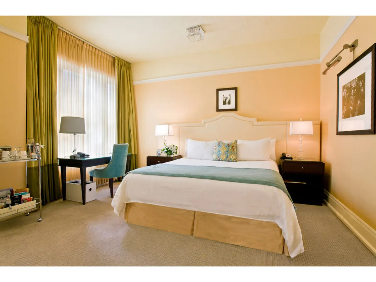 A welcoming bedroom scene with a plush king-size bed dressed in elegant linens and decorative pillows. Oversized windows draped in sheer curtains bathe the room in natural light. Beside the bed sits a stylish nightstand and a sophisticated desk with a comfortable chair, offering both a space for productivity and relaxation. The mood is peaceful and refined, capturing guests reading by the window, working at the desk, or simply unwinding on the bed.