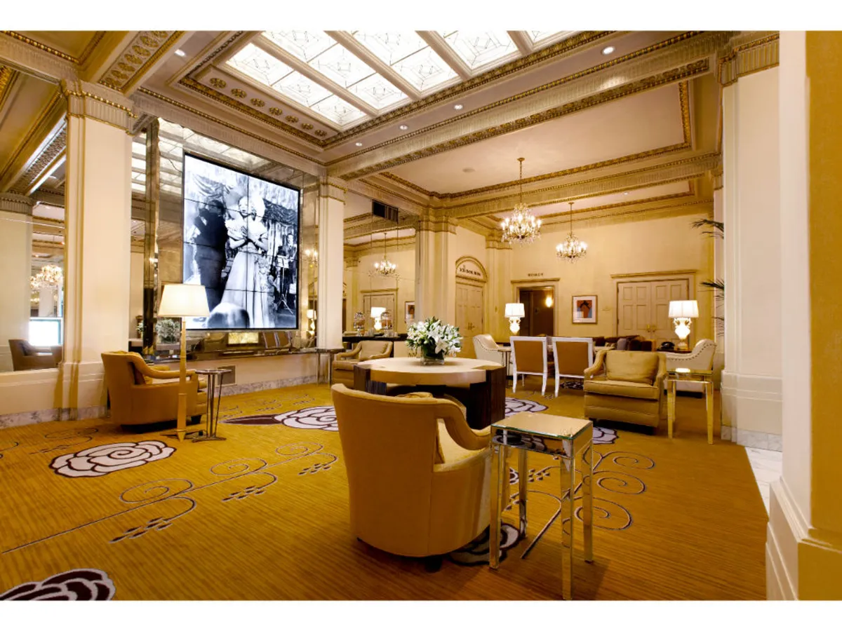 A sophisticated screening or event space with a large screen displaying historical imagery, ornate chairs arranged around a round table with a floral centerpiece, and luxurious chandeliers overhead. The decorative carpet adds to the refined setting.