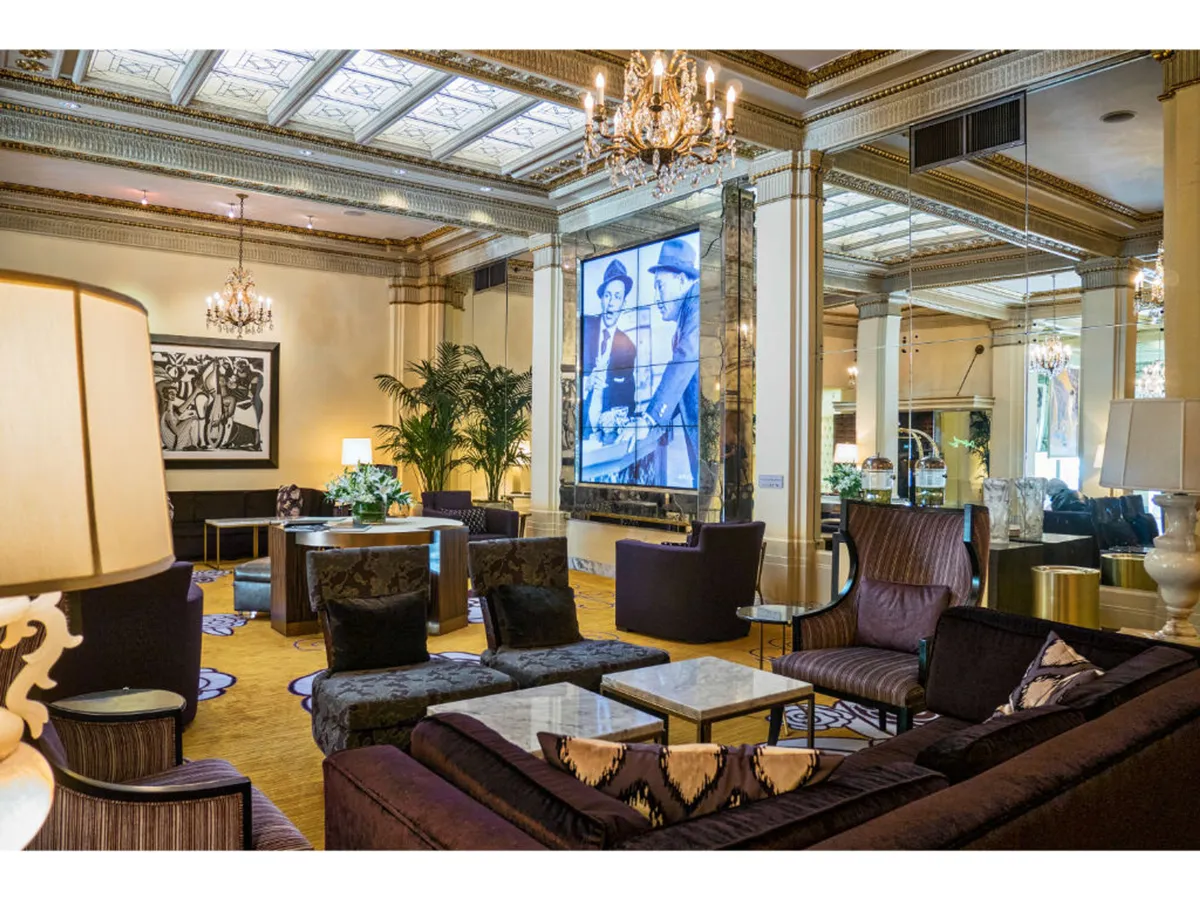 A refined interior lounge features a chandelier, large screen display, comfortable sofa arrangements, and art adorning the walls. The space is ideal for informal meetings, relaxation after a long day, or even hosting small gatherings for clients.